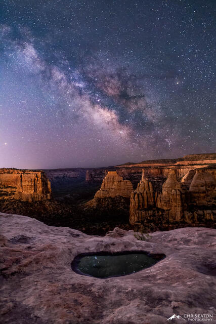 The rising Milky Way is reflected in a pothole of water along the cliff rim above Monument and Wedding Canyons.