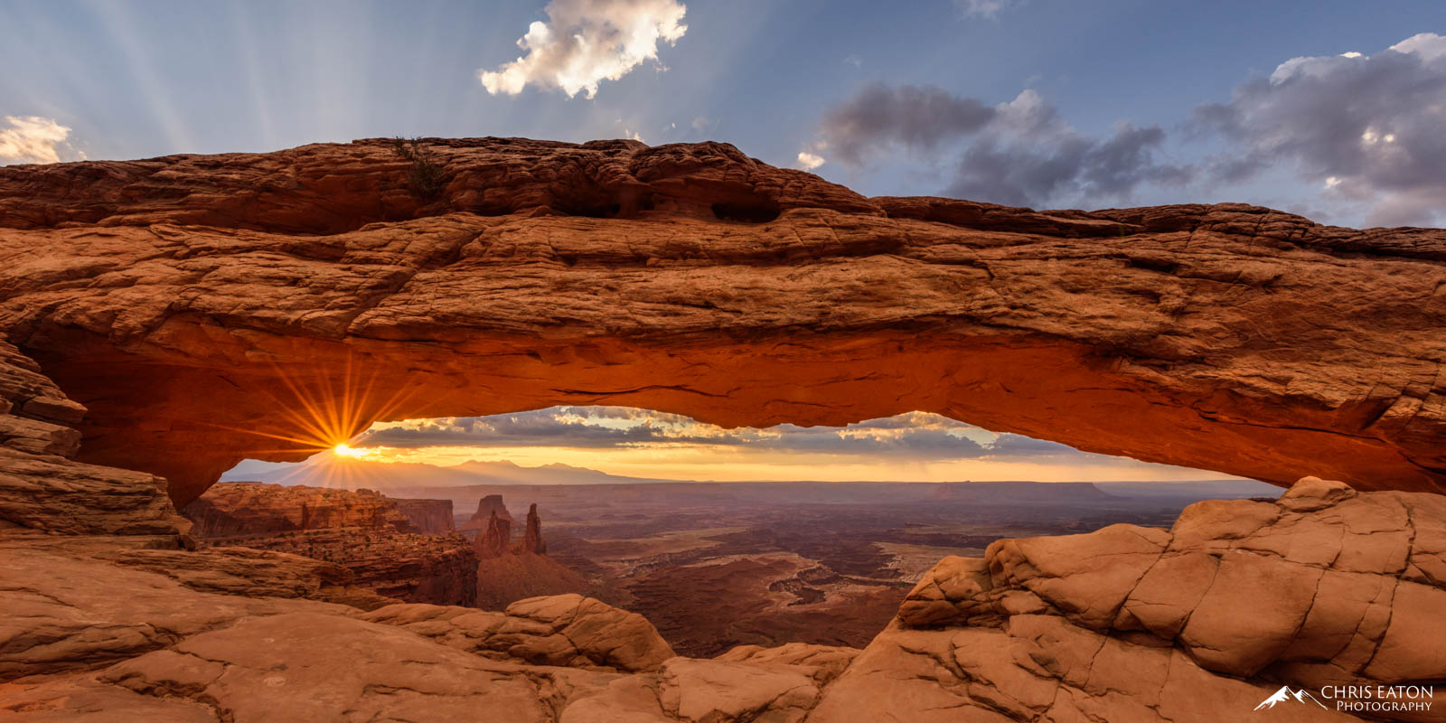 The rising sun bursts through a low layer of clouds over the La Sal Mountains, painting the underside of the arch with vibrant...