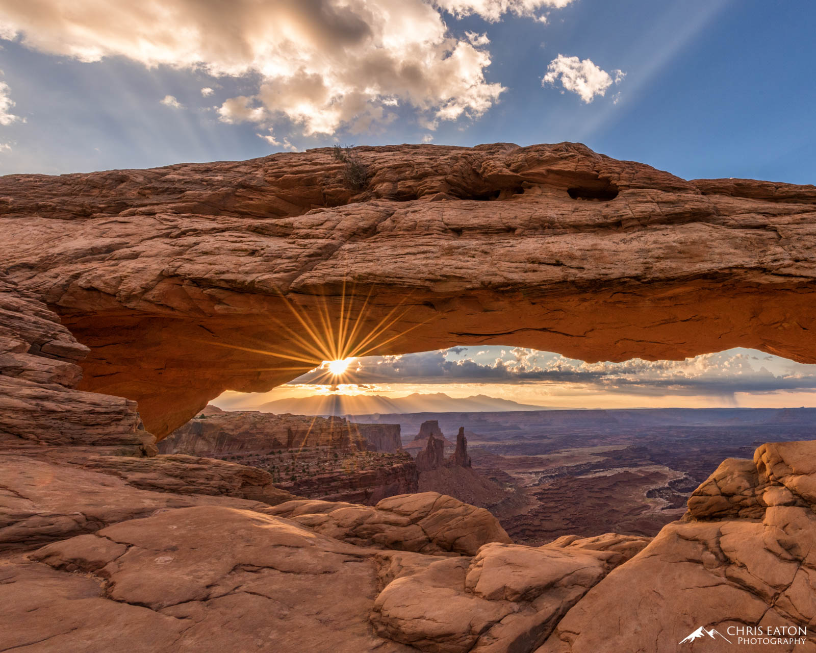 The sun bursts through a low layer of clouds over the La Sal Mountains, painting the underside of the arch with vibrant reflected...